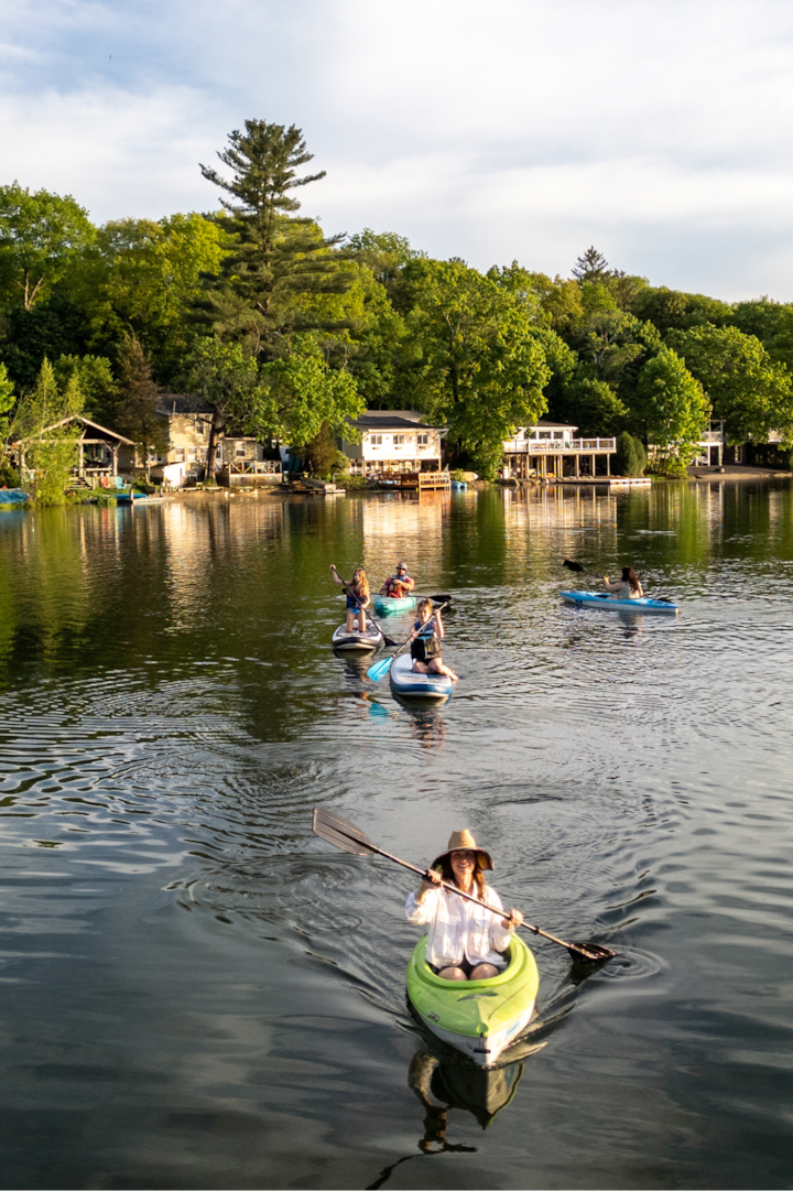 Un groupe de personnes fait du paddle et du kayak sur un lac paisible. Des maisons pittoresques se dressent à l'arrière-plan, près de la rive et nichées dans un écrin de verdure.