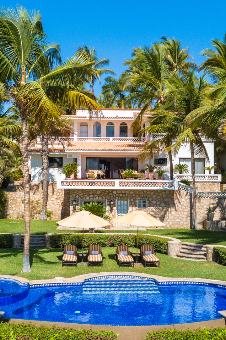 Une villa de luxe avec des tuiles en terre cuite, des palmiers luxuriants, un jardin paysager et une piscine d'un bleu étincelant avec des sièges au bord de l'eau.