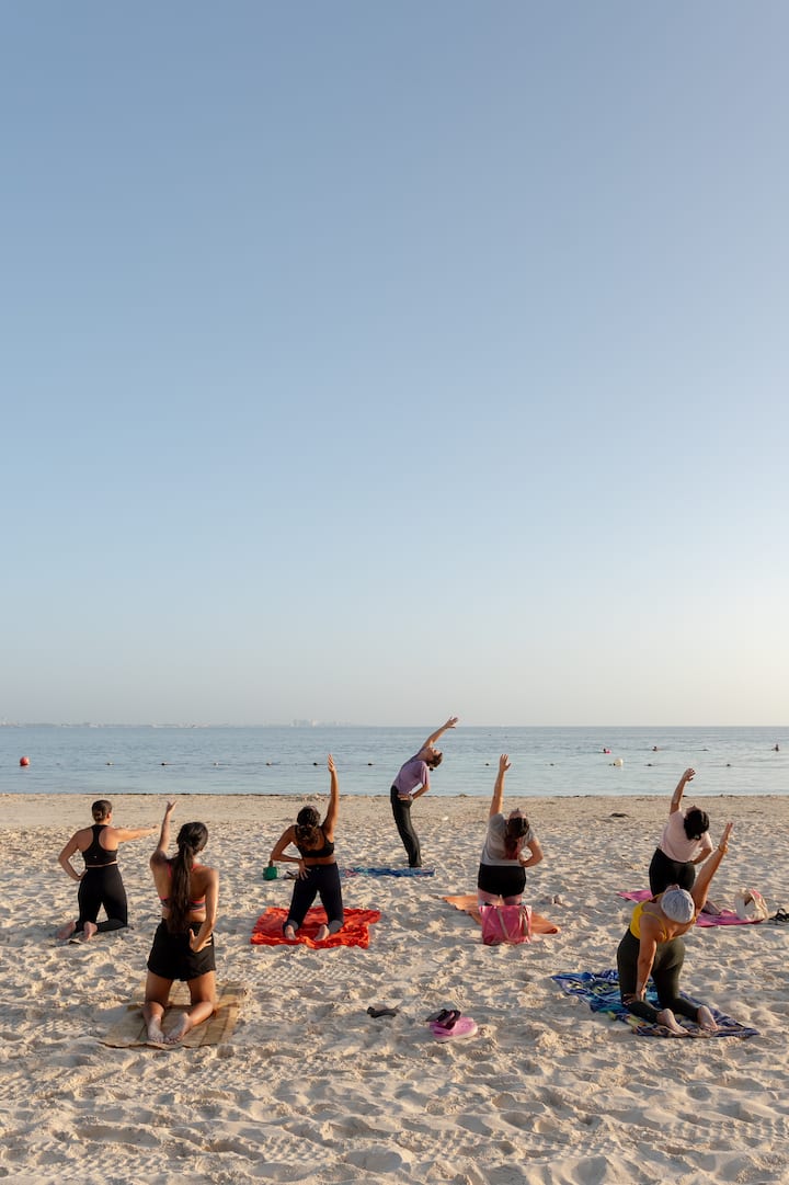 Un groupe de personnes pratiquant le yoga sur une plage de sable fin. Un océan calme sous un ciel bleu limpide se trouve à l'arrière-plan.