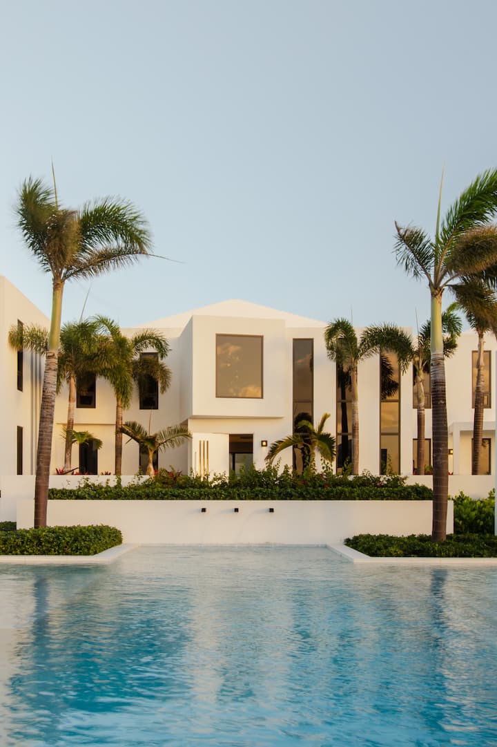 Une villa blanche et moderne entourée de palmiers, avec de grandes fenêtres et une piscine au premier plan, sous un ciel dégagé.
