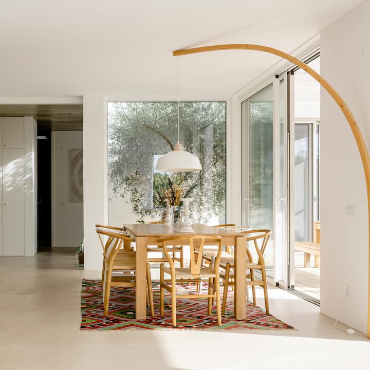 Un espace repas lumineux, avec une table et des chaises en bois, un tapis tissé coloré et de grandes portes vitrées donnant sur un patio et de la verdure.