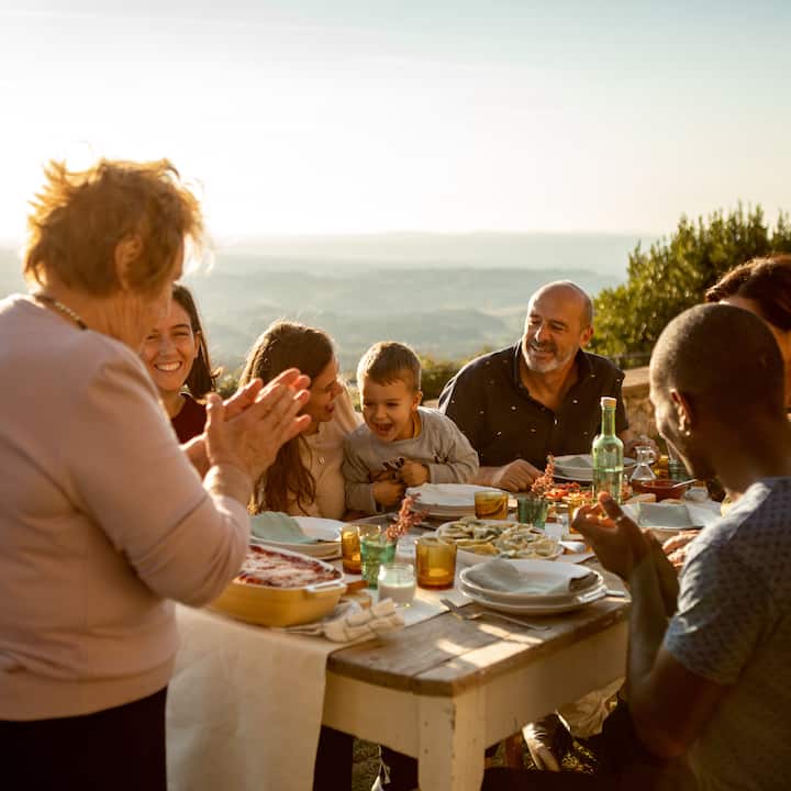 Un groupe de personnes prenant un repas en extérieur au coucher du soleil. Elles sont assises autour d'une table rustique sur laquelle sont posées des boissons et des assiettes contenant de la nourriture, le tout surplombant un paysage pittoresque.