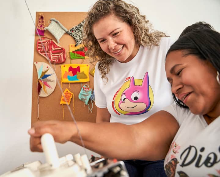 Dos personas están reunidas en torno a una máquina de coser en una habitación con obras de arte de colores en la pared.