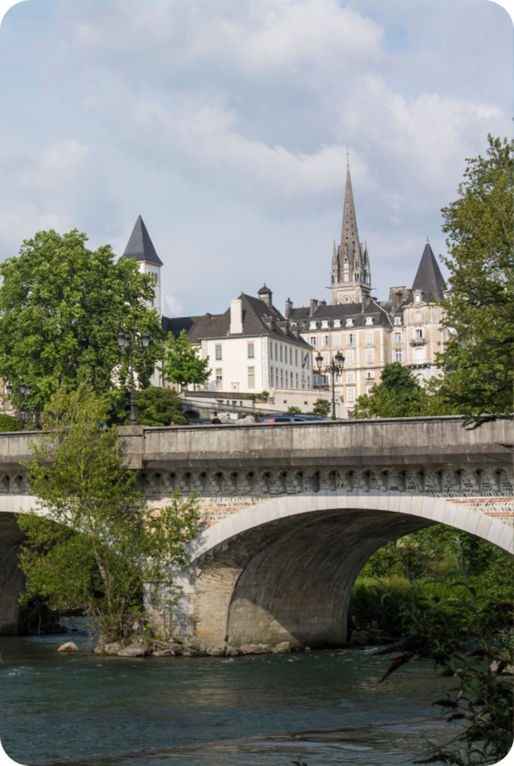 Un pont en pierre enjambe une rivière, avec en arrière-plan des bâtiments historiques. Texte superposé : « Étape 6 – Pau », avec une flèche vers la droite.