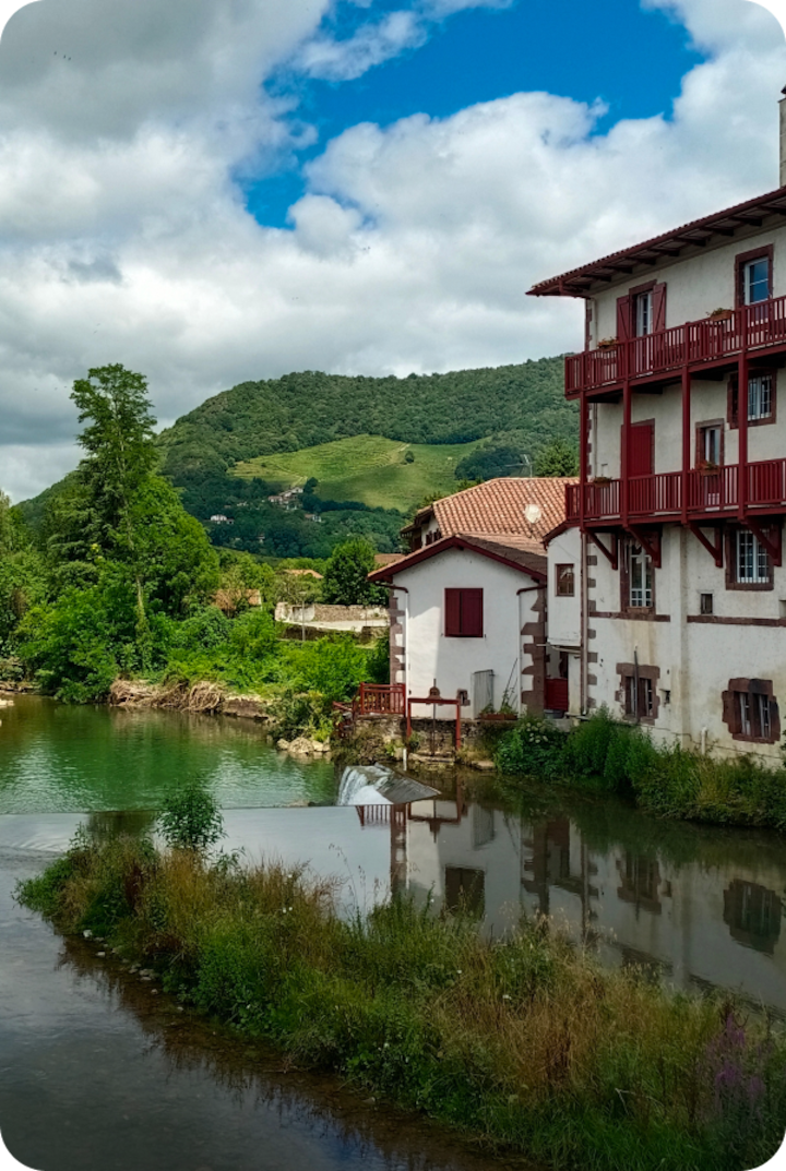 Des maisons blanches aux volets et balcons rouges bordent une petite rivière. Texte superposé : « Étape 7 – Hagetmau », avec une flèche vers la droite.