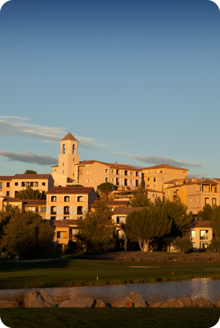 Un village à flanc de colline au coucher du soleil, avec des bâtiments aux teintes pastel et un clocher dominant. Texte superposé : « Étape 9 – Malemort », avec une flèche vers la droite.
