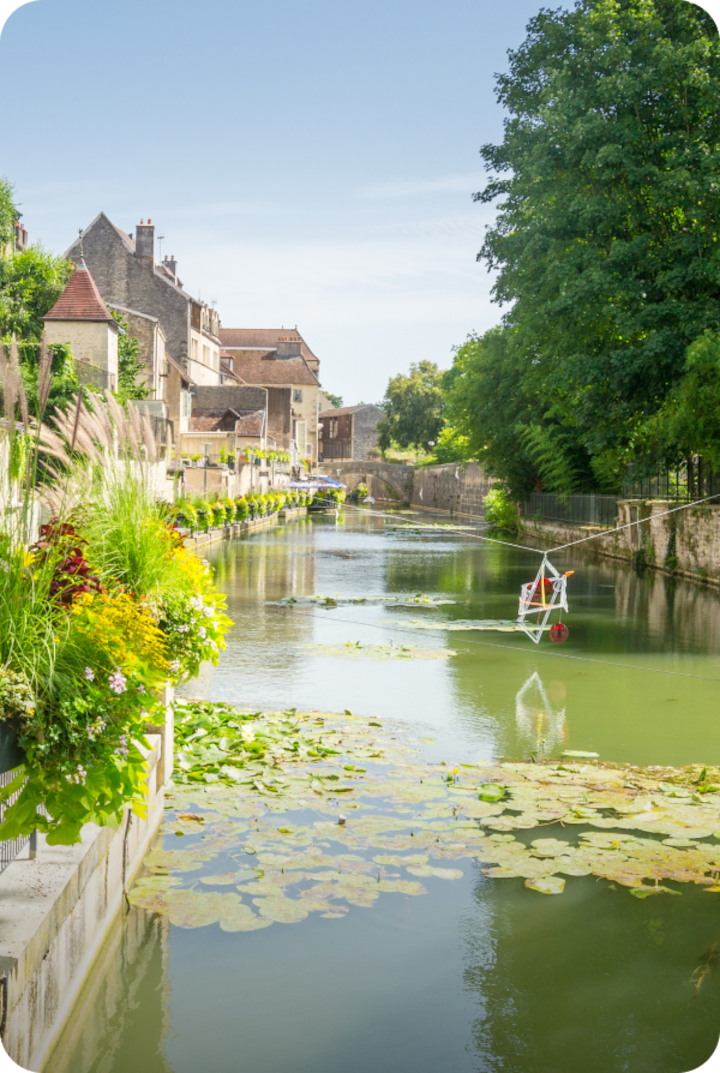 Un canal paisible bordé de bâtiments, de verdure et de fleurs colorées. Texte superposé : « Étape 13 – Dole », avec une flèche vers la droite.