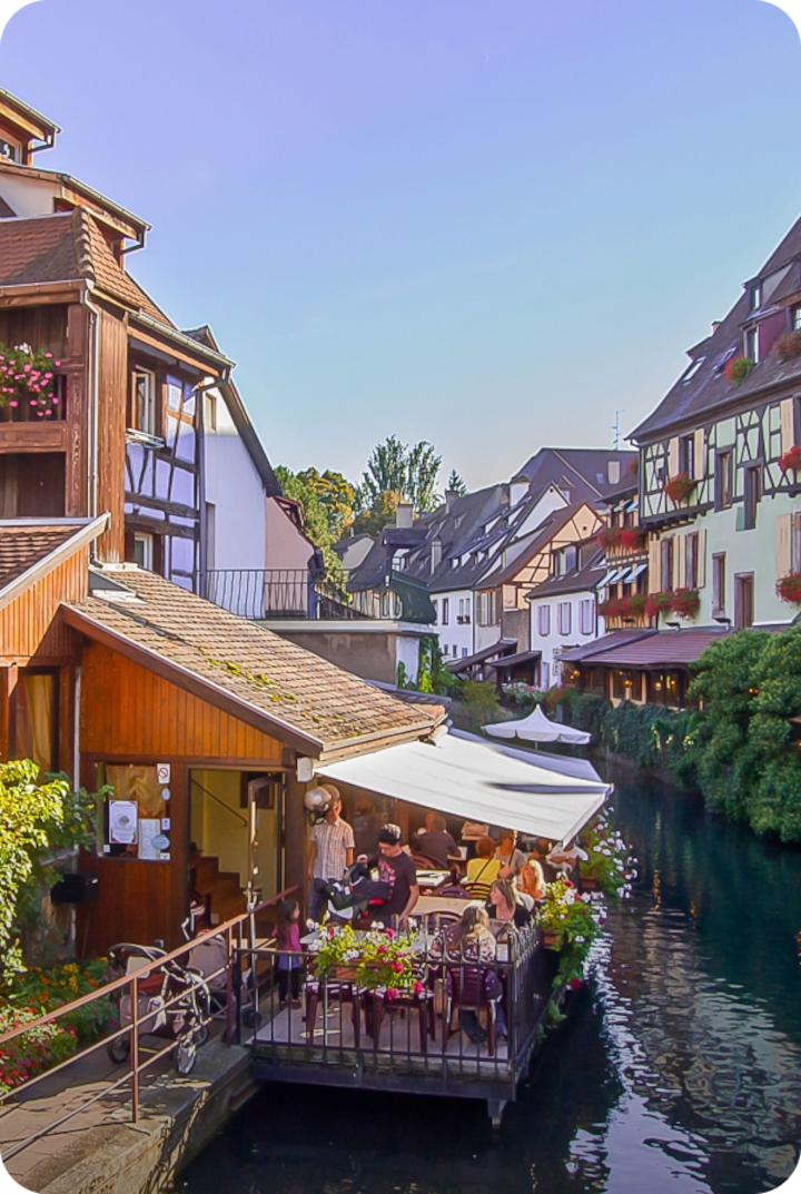 Un canal bordé de maisons à colombages, avec des jardinières fleuries aux balcons. Texte superposé : « Étape 14 – Mulhouse », avec une flèche vers la droite.