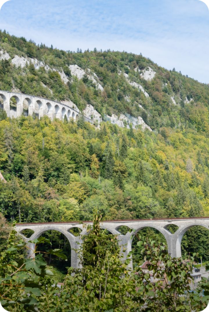Un paysage verdoyant avec deux viaducs ferroviaires en pierre aux arches multiples. Texte superposé : « Étape 15 – Champagnole », avec une flèche vers la droite.