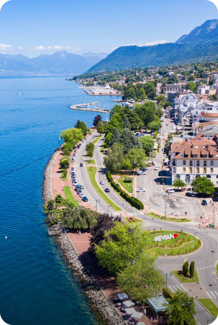 Vue aérienne d'une ville au bord d'un lac, avec une promenade bordée d'arbres et de routes. Texte superposé : « Étape 16 – Évian-les-Bains », avec une flèche vers la droite.