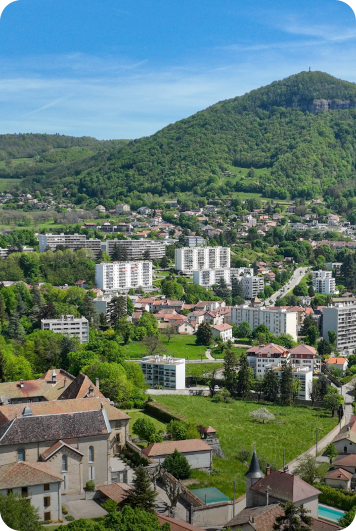 Vue aérienne de Voiron nichée au cœur de collines verdoyantes. Texte superposé : « Étape 18 – Voiron », avec une flèche vers la droite.