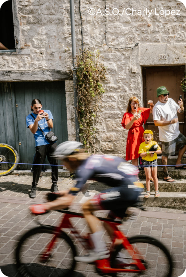 Des personnes sur les marches d'une maison rustique regardent passer deux cyclistes professionnels roulant à toute vitesse. Texte superposé : « Étape 20 – Le Bourg-d'Oisans », avec une flèche vers la droite.