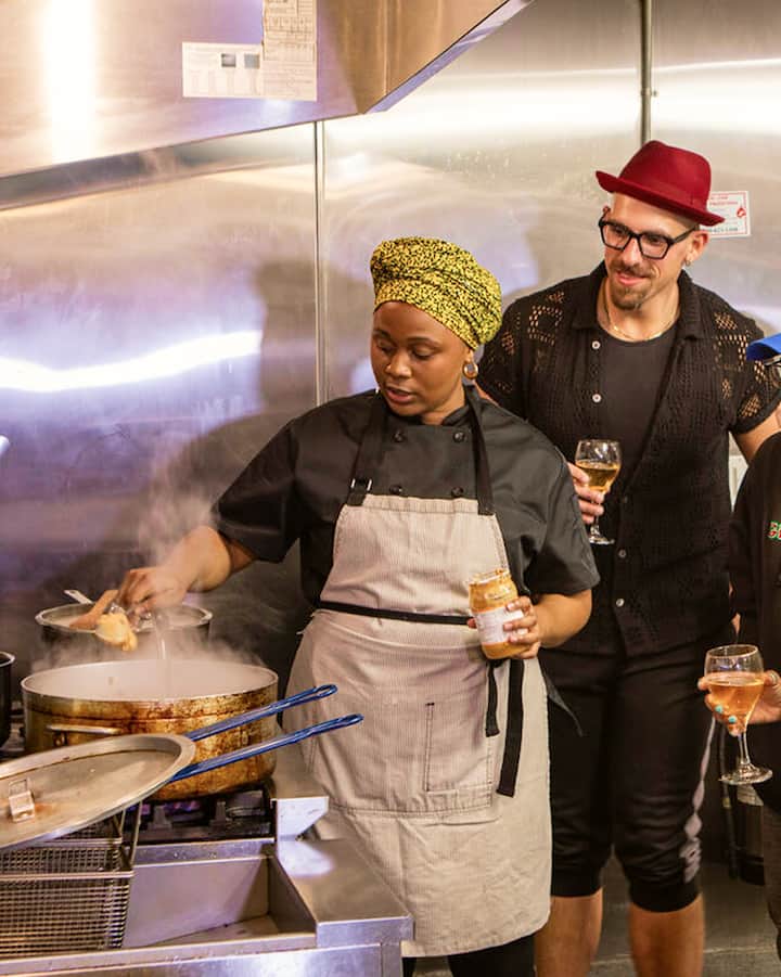 People with wine glasses in hand gather around a chef as she cooks in a professional kitchen