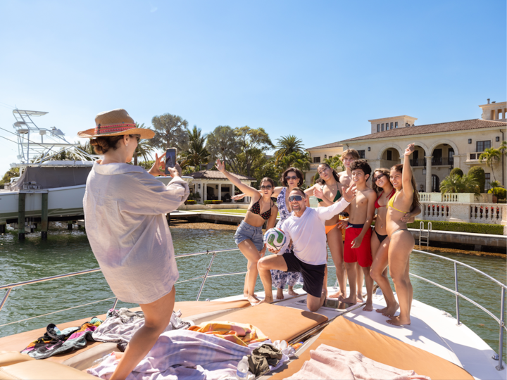 Friends in swimwear pose on a sunny yacht while someone takes their photo, celebrating together on the water.