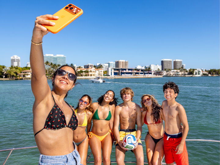 Friends in swimwear take a boat selfie with a soccer ball, celebrating the tournament on sunny waterfront city waters.