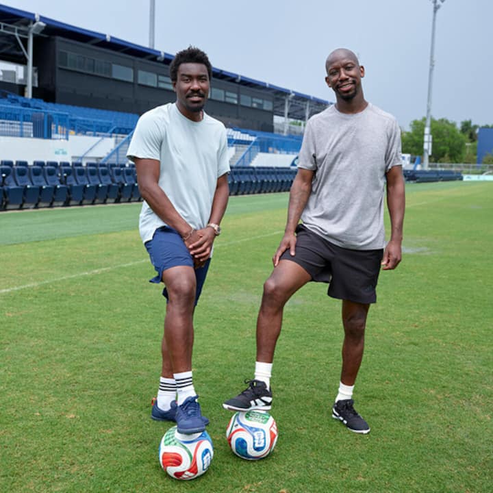 Two soccer players stand on the grass at Miami Stadium, each resting a foot on a FIFA World Cup 26 ball.