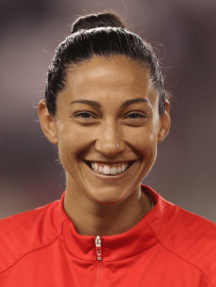 Un jugador de fútbol con una chamarra roja de calentamiento está de pie en un estadio; su rostro está desenfocado por motivos de privacidad.