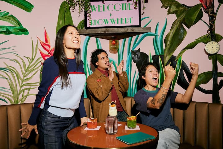 Amigos celebrando juntos en un bar colorido, brindando y vitoreando alrededor de una mesa en pleno día de partido.
