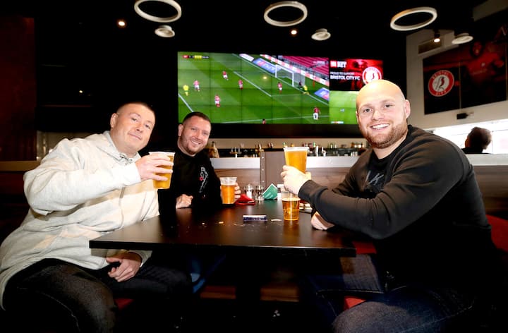 Un grupo de aficionados sentados juntos en un bar, brindando con cervezas mientras ven un partido de fútbol en una televisión de pantalla grande.