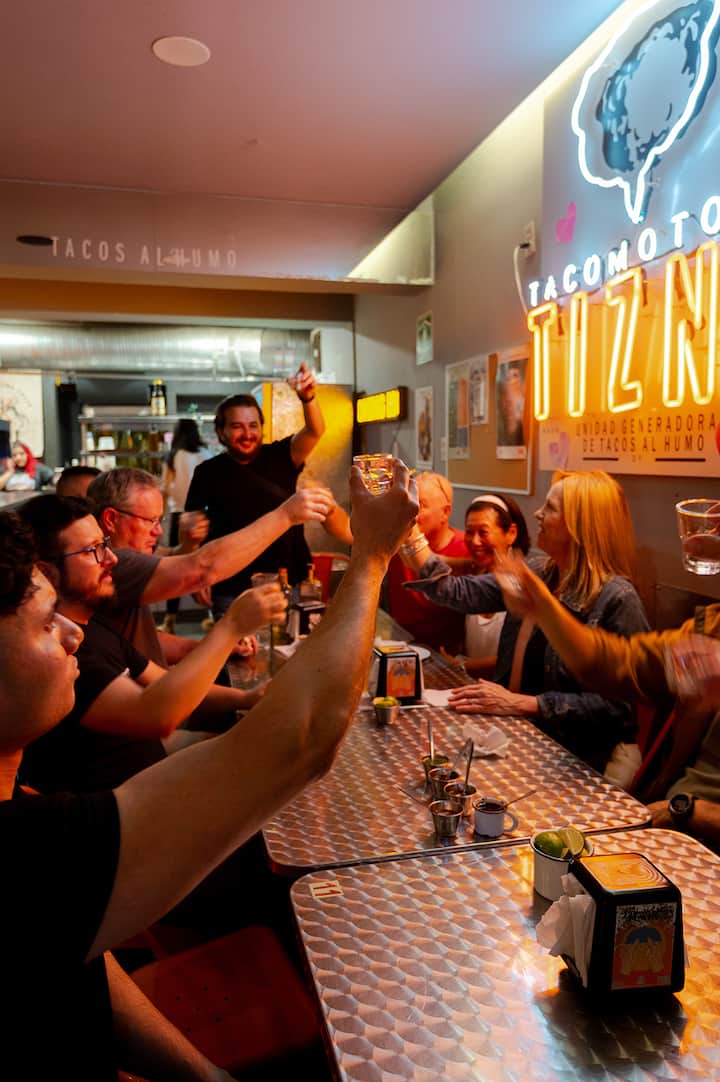 Friends raise tequila shots around a metal table at a lively taco bar in Mexico City.