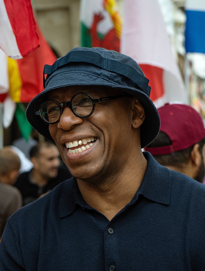 Gros plan d'Ian Wright en tenue décontractée, il se trouve au milieu d'une fête de rue où flottent des drapeaux de différents pays.