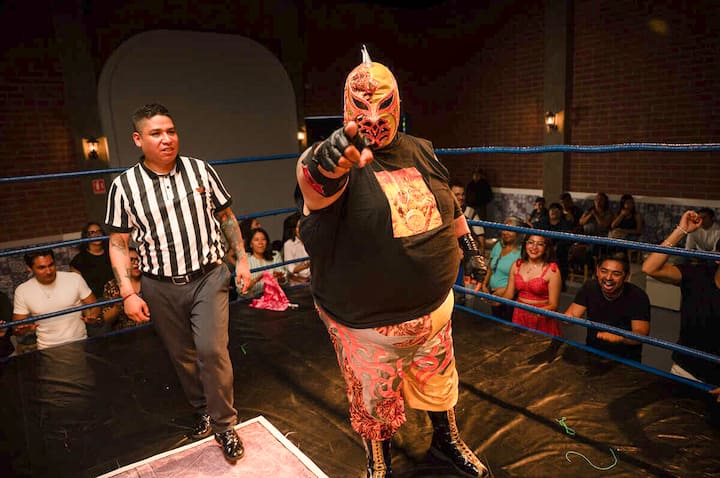 Lucha libre wrestler in a mask points at the camera in a packed Mexico City arena, referee and crowd behind him.