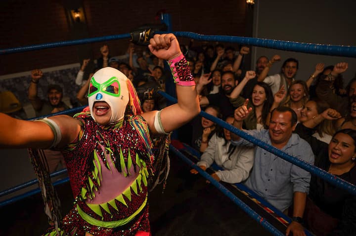 Masked luchador in a colorful costume celebrates in a Mexico City ring as fans cheer with raised fists.