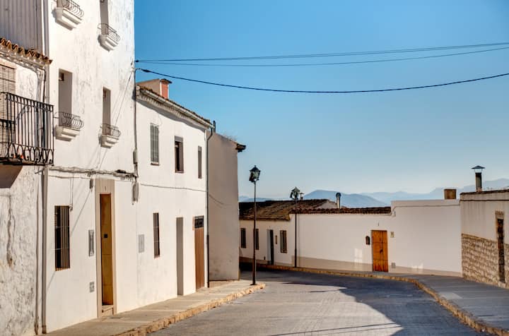 Quiet sunlit street in a rural Spanish village with whitewashed houses and distant mountains.