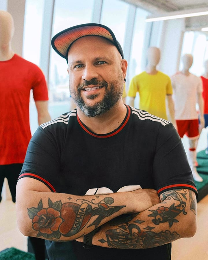 Un homme souriant, les bras croisés, pose devant un éventail de maillots de soccer de différentes couleurs.