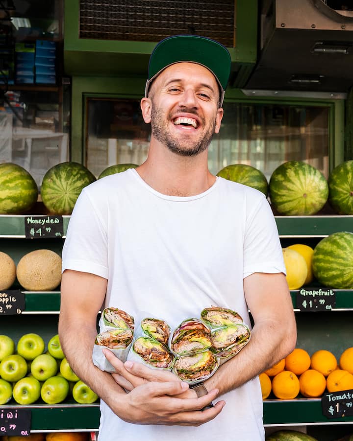 Un homme souriant pose devant un kiosque de fruits et légumes en tenant quatre sandwichs alléchants dans ses bras.