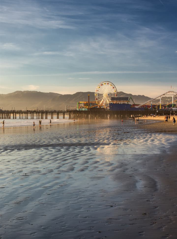 Sunset view of Santa Monica Pier with its lit Ferris wheel reflected on the wet beach shoreline - view more listings in LA.
