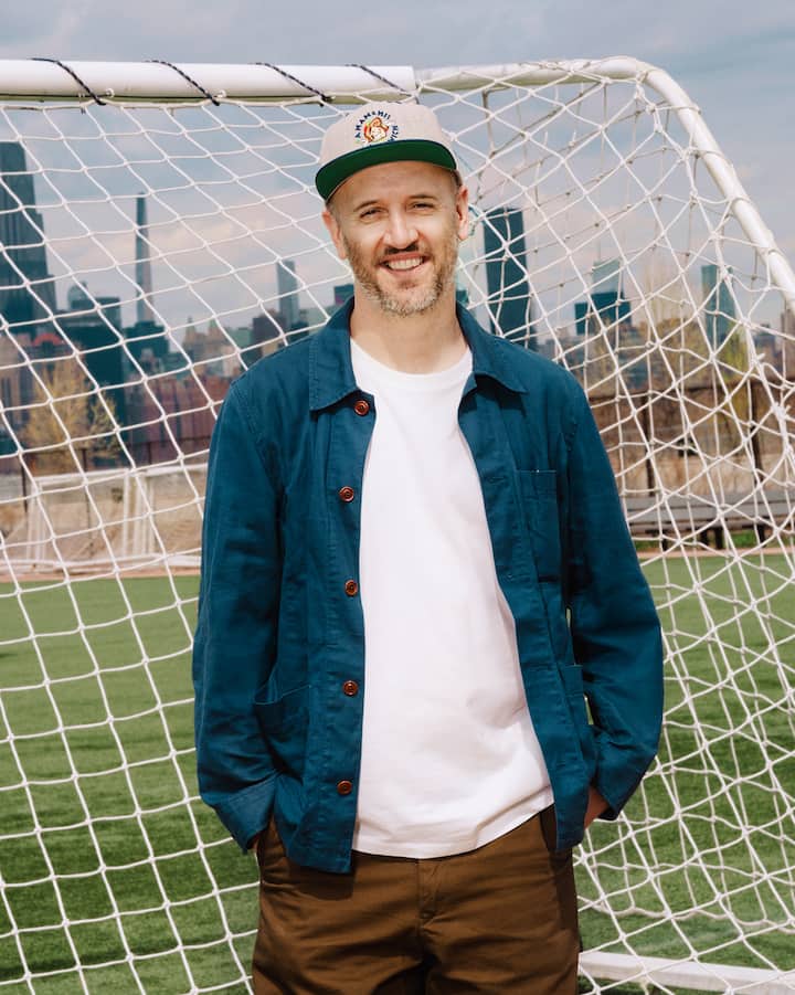 Un homme portant un manteau bleu et une casquette de baseball grise fait un sourire à la caméra devant un but.