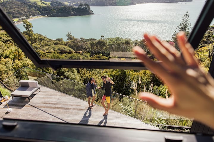 Friends wave up at an open window from a sunny deck overlooking a lake and lush green hills