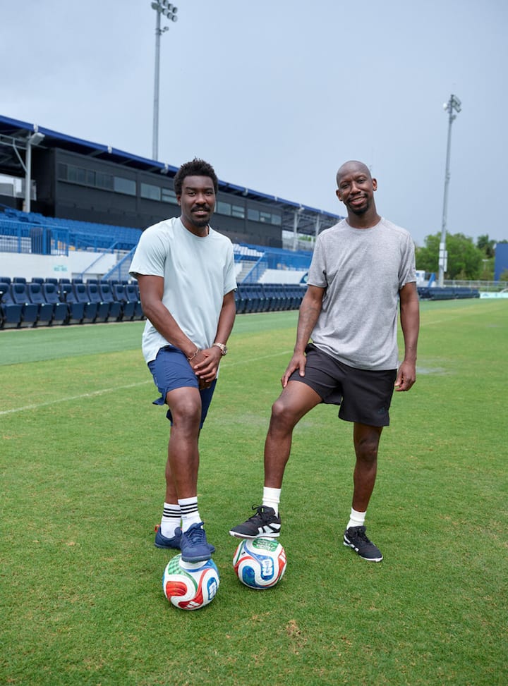 Dos amigos están de pie en un campo de fútbol de Miami. Cada uno apoya un pie en un colorido balón de la Copa Mundial de la FIFA.