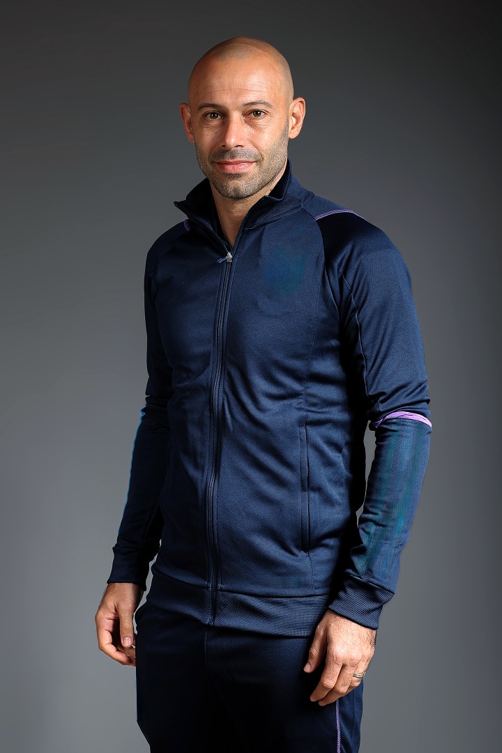 Una persona con un chándal azul marino oscuro, de pie frente a un fondo gris, con un estilo que recuerda a la equipación oficial de los aficionados de la FIFA.
