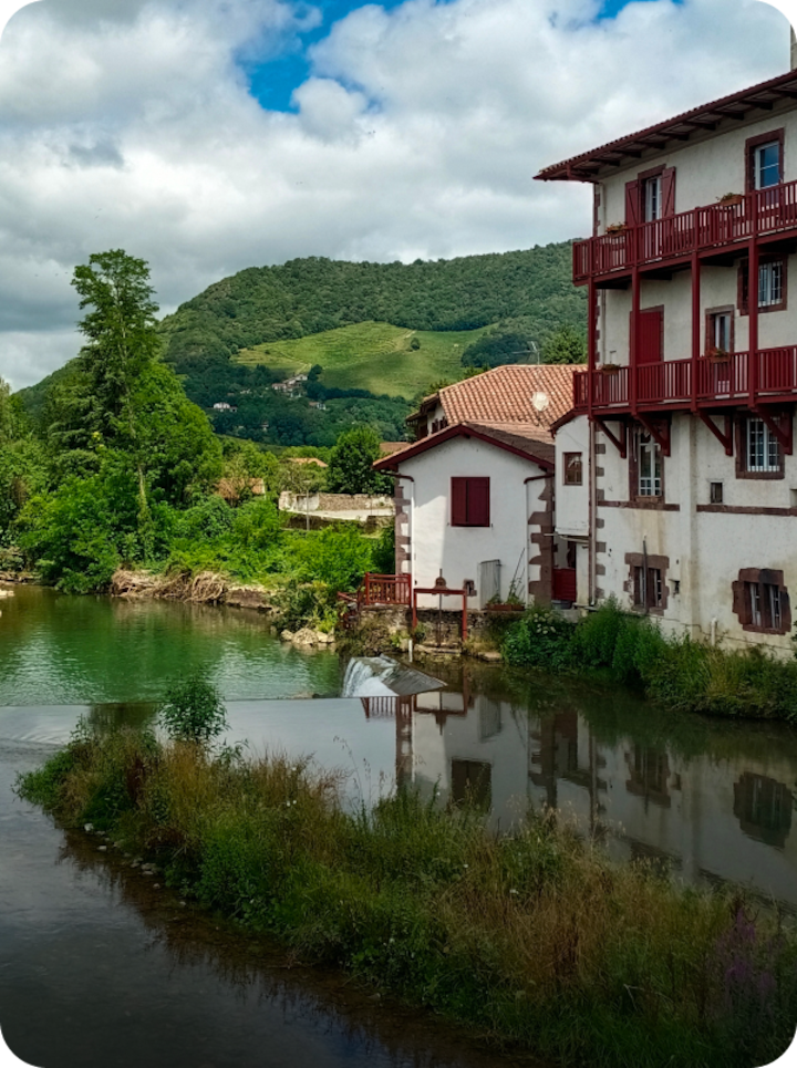 Des maisons blanches aux volets et balcons rouges bordent une petite rivière. Texte superposé : « Étape 7 Hagetmau », avec une flèche vers la droite.