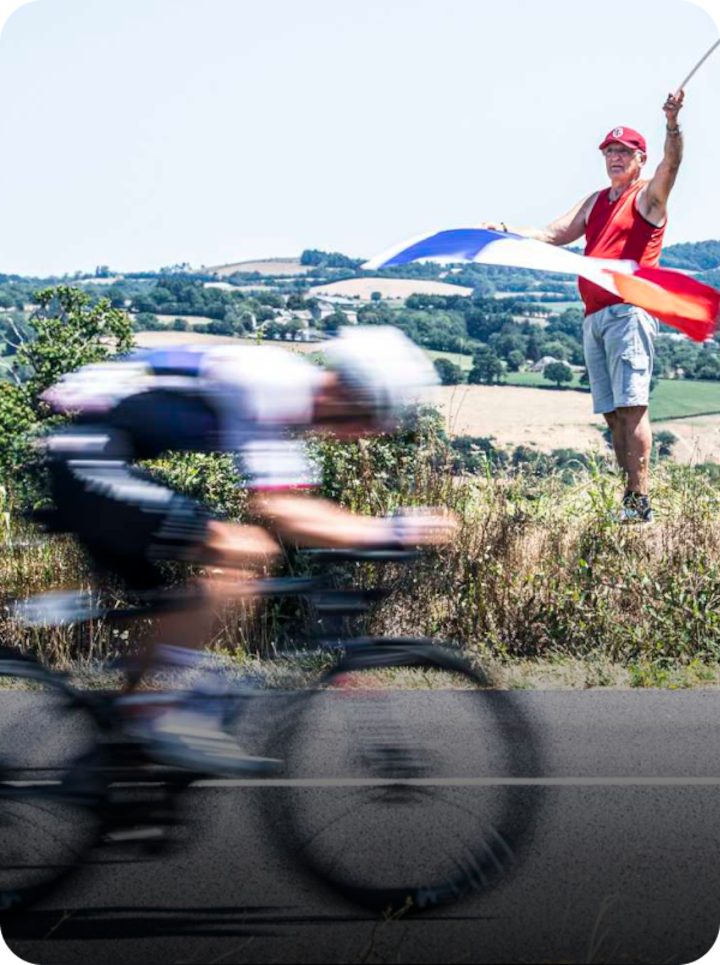 Un cycliste roule sur une route de campagne, tandis qu'une personne se tient sur l'accotement herbeux et agite un grand drapeau français. Texte superposé : « Étape 12 Magny-Cours », avec une flèche vers la droite.