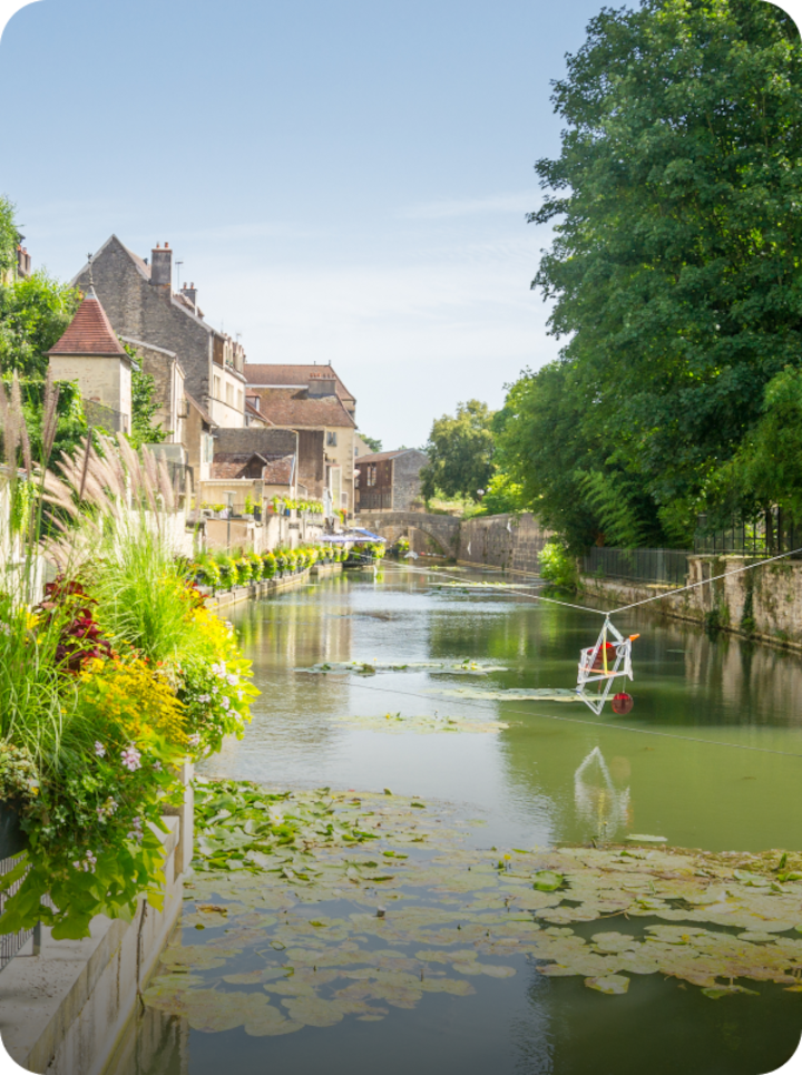 Un canal bucolique bordé de bâtiments, de végétation et de fleurs multicolores. Texte superposé : « Étape 13 Dole », avec une flèche vers la droite.