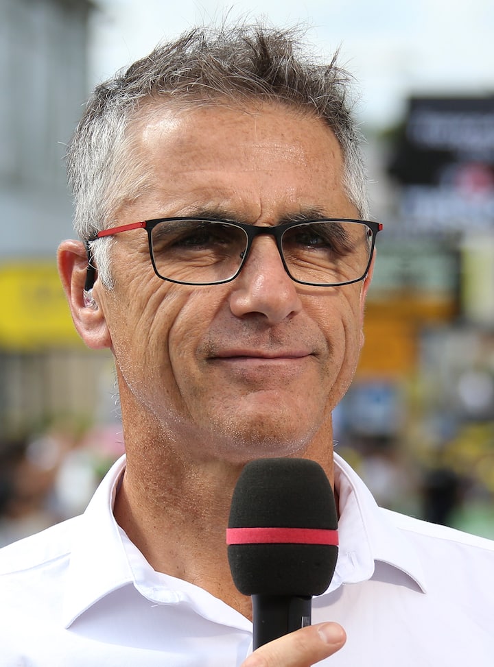 Commentateur sportif tenant un micro lors d'un événement, avec la foule de la course floutée à l'arrière-plan.
