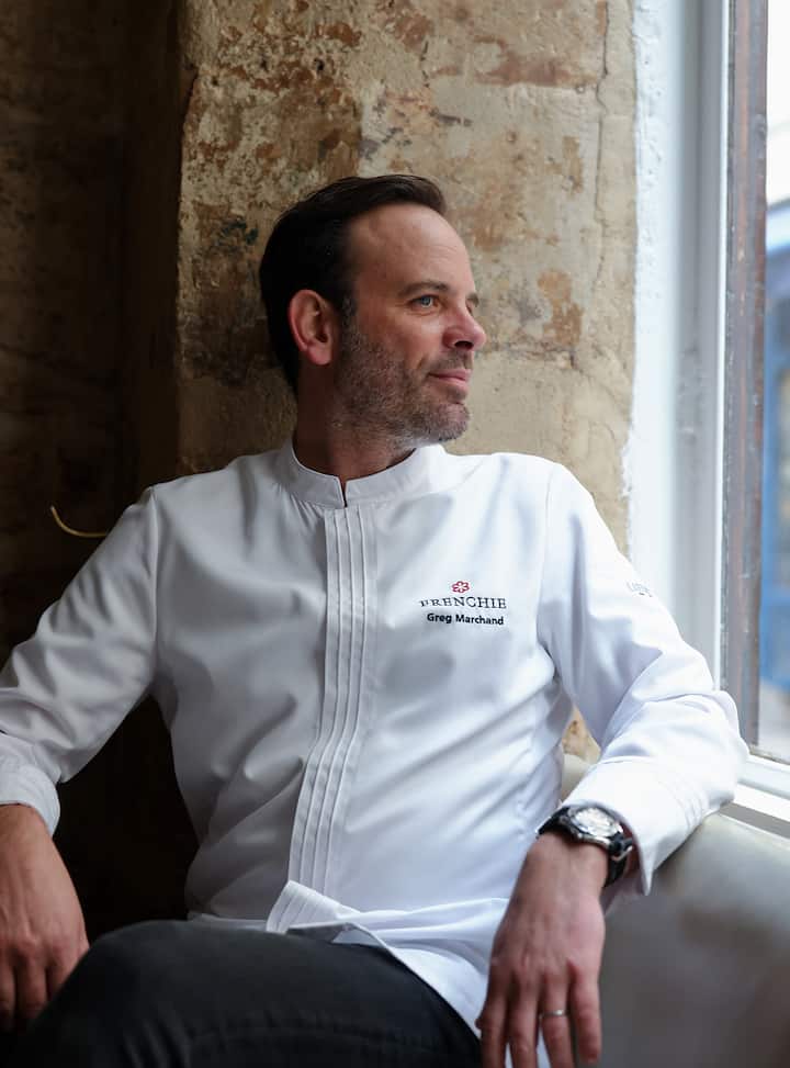Un chef portant une veste blanche siglée Frenchie et brodée avec le nom Greg Marchand est assis devant une fenêtre.