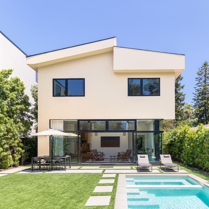 Une maison moderne à deux niveaux donnant sur un jardin avec terrasse, des chaises longues et une petite piscine.