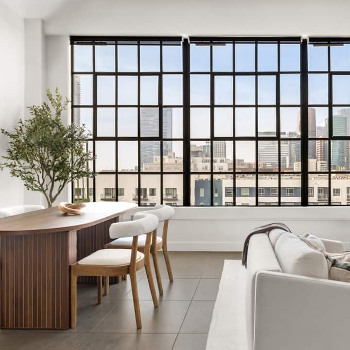 Un salon ensoleillé dans un appartement moderne, doté de fenêtres allant du sol au plafond et offrant une vue sur le panorama urbain de Los Angeles.