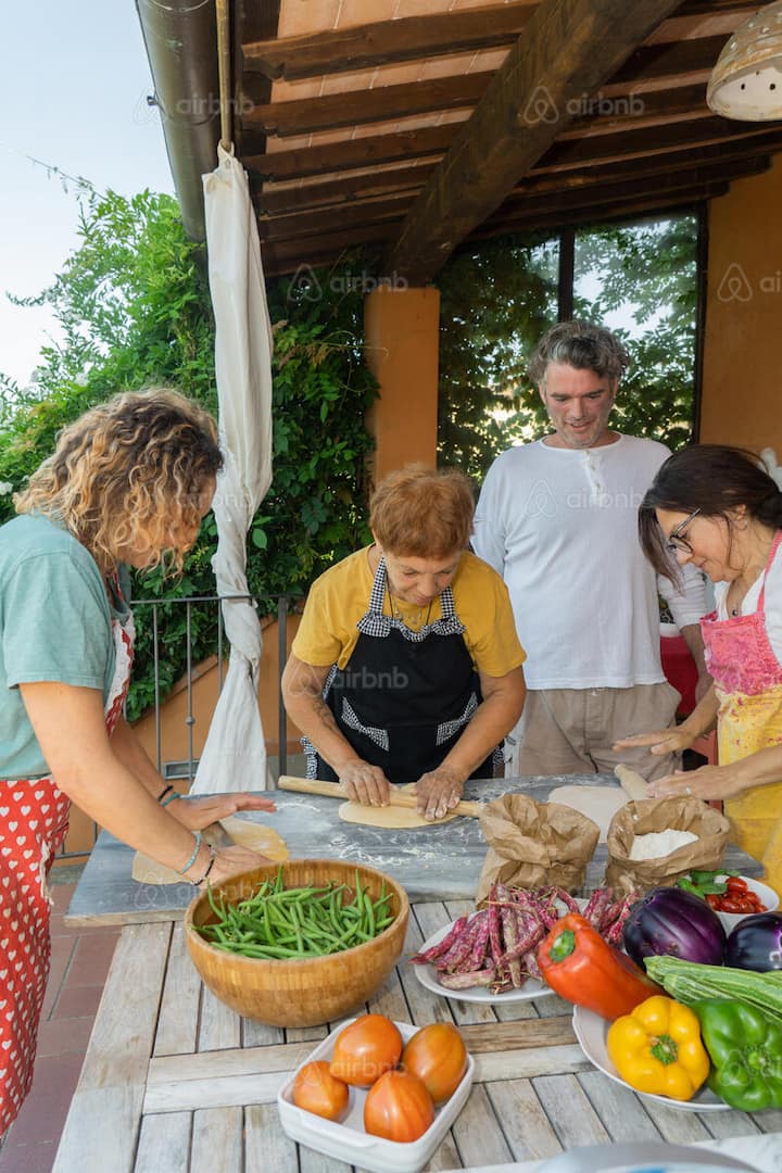 Guests gather on a terrace for a hands-on cooking class, rolling dough beside colorful fresh vegetables.