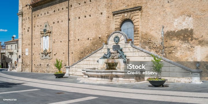 Piazza di un piccolo borgo italiano con antica fontana in pietra davanti a un edificio storico.