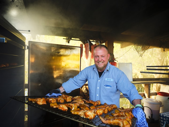 Outdoor host cooking a full rack of smoked fish, steam rising from the open metal smoker