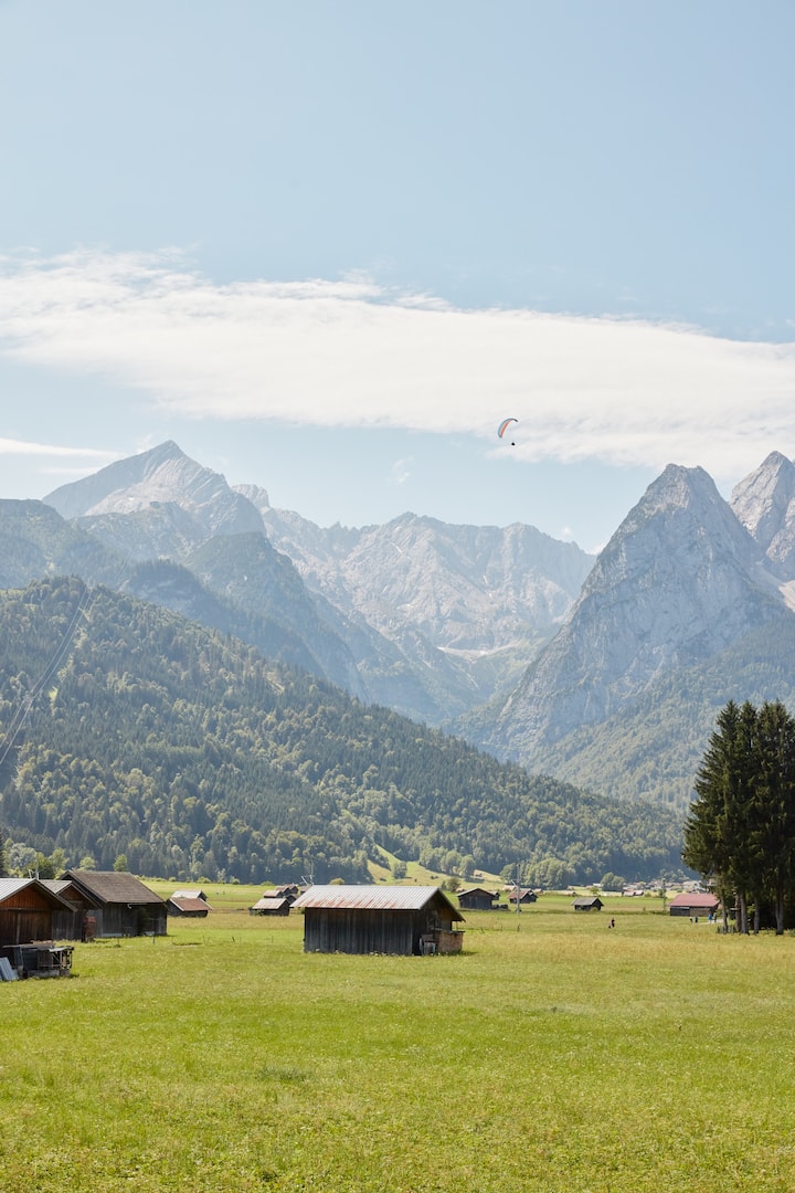 Wide green meadow with small barns, forested German Alps, and a lone paraglider in the sky