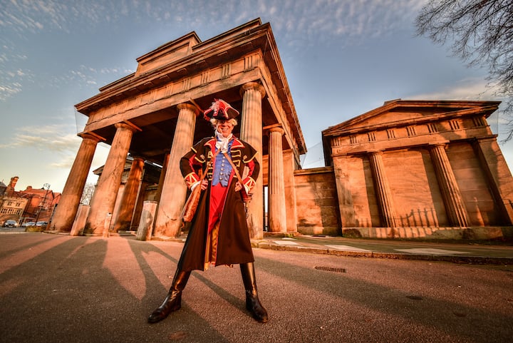 Person in colorful British town crier costume posing proudly before grand stone columns at sunset.