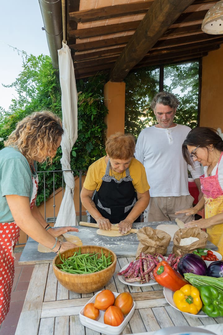 Group of guests making fresh pasta together on a shaded patio, surrounded by colorful local vegetables.