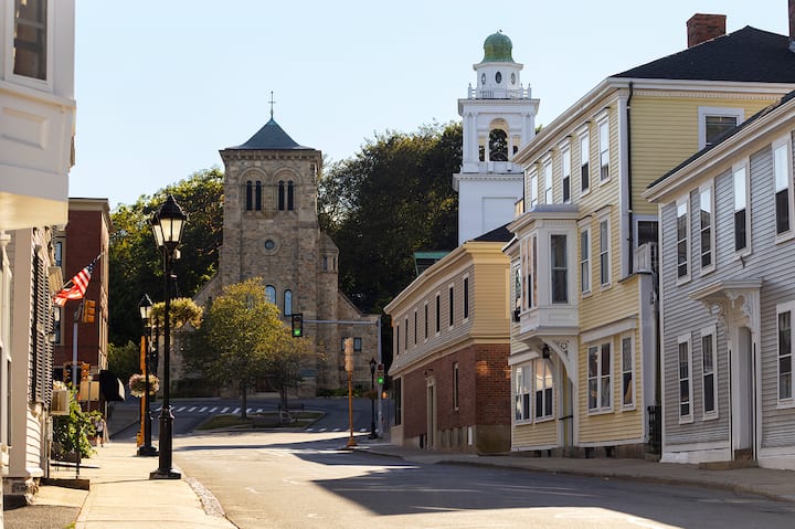 A photo of a quiet street in Massachusetts, with a hanging American flag and historical building in the background.