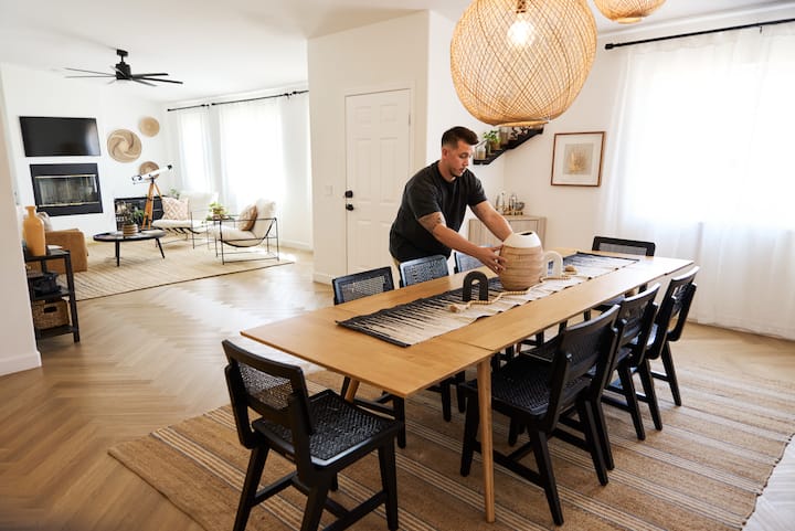 Cleaner tidies a bright Airbnb dining room, setting a woven vase on a long wooden table.